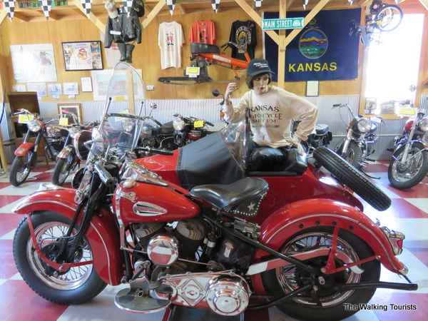 An old motorcycle centered in front of other ones and shirts on a wall