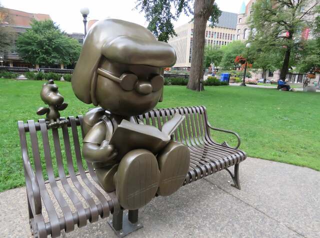 Marcie reading a book while sitting on a bench with Woodstock the bord behind her