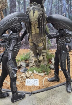 Two Alien creatures look at a Predator they have captured at a display